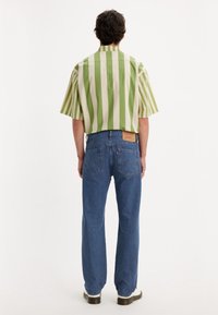 Kortärmad skjorta med gröna och krämfärgade vertikala ränder, ihop med ljusblå jeans och vita skor. Bakifrån syns en etikett på jeansen.