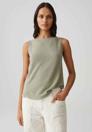 Frau mit langen dunklen Haaren, die ein ärmelloses hellgrünes Oberteil und weiße Hose trägt, mit einer Hand in der Tasche vor grauem Hintergrund.