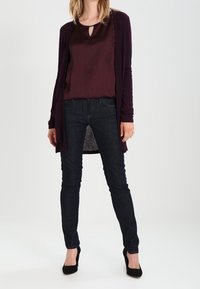 Kobieta w ciemnofioletowej bluzce i kardiganie, ciemnych, dopasowanych dżinsach oraz czarnych szpilkach stoi na tle jednolitego tła.