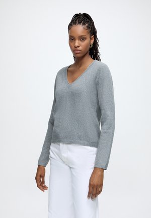 Mujer con el cabello trenzado lleva un suéter gris con cuello en V y pantalones blancos, de pie frente a un fondo claro y liso con expresión neutral.