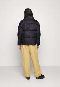 Black padded jacket with a high collar and subtle branding, paired with beige cargo pants and black platform shoes. Smooth texture, oversized fit.