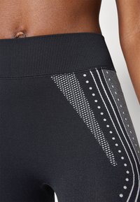 Legginguri atletice negre, cu o talie largă din material canelat. Detalii laterale cuprind diverse modele texturate în alb, inclusiv puncte și linii.