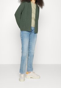 Green knitted cardigan, short sleeves, open front, worn over a light green T-shirt and paired with light blue jeans and beige sneakers.