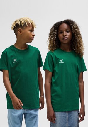 Zwei junge Menschen, die grüne Hummel-T-Shirts tragen, einer schaut seitlich, der andere blickt nach vorne, beide mit lässigen Frisuren und Jeansbekleidung.