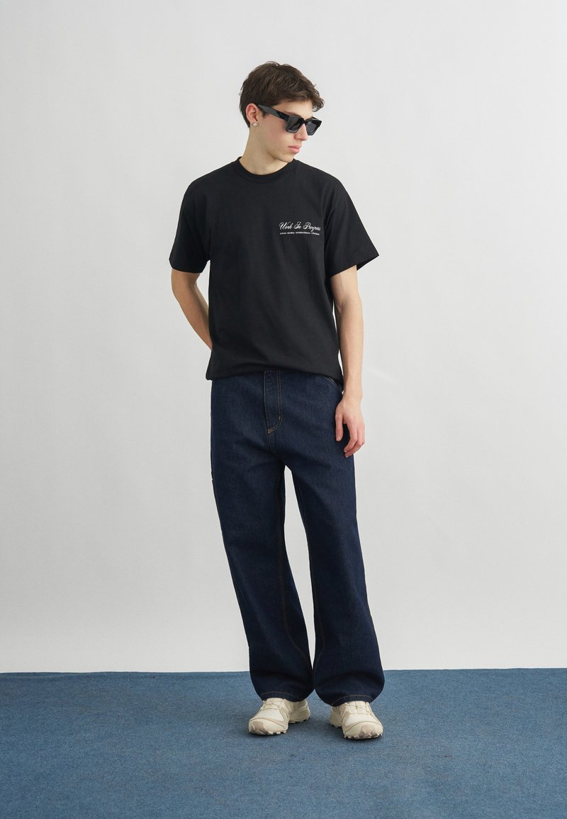 Giovane uomo con occhiali da sole neri, t-shirt grafica nera, jeans blu scuro e sneakers bianche in piedi su un tappeto blu contro una parete bianca.