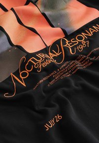 Tissu noir plié avec un texte orange indiquant "Nocturnal Resonance Festival 1987" et "26 juillet" accompagné de formes abstraites orange et grises.