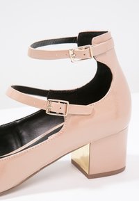 Chaussure à talon bas en cuir verni rose avec double bride à la cheville et boucles carrées dorées, dotée d'un talon bloc avec un accent métallique doré.