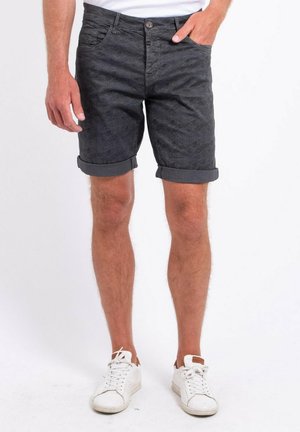 Homme portant un short foncé à motifs retroussé et des baskets blanches, debout avec une main dans la poche devant un fond uni.