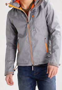 Giacca grigia con accenti arancioni, dotata di cappuccio, multiple tasche con zip e una finitura testurizzata. Vestibilità casual, indossata sopra dei jeans.