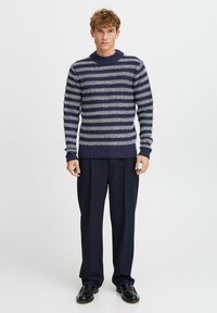 Maglione a maglia blu navy con strisce orizzontali bianche, colletto a giro, e polsini e orlo a costine, abbinato a pantaloni scuri e scarpe nere.