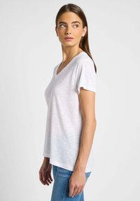 Camiseta blanca de algodón con cuello en V, de manga corta, corte holgado y textura suave. Presenta un patrón jaspeado sutil. Jeans visibles por debajo.