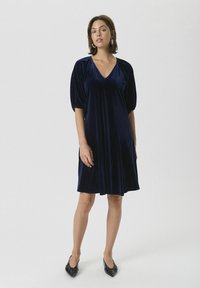 Rochie din catifea bleumarin, cu decolteu în V, mâneci scurte bufante și fustă evazată. Este purtată cu pantofi cu vârf ascuțit negri.