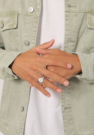 Anello d'argento con un design geometrico, indossato su una mano, abbinato a una giacca di jeans verde chiaro e una t-shirt bianca. Texture liscia, stile informale.