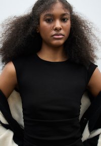 Musta hihaton toppi, joka on valmistettu sileästä kankaasta ja jossa on vartalonmyötäinen siluetti. Mallilla on hartioillaan kontrastivärinen mustavalkoinen takki.