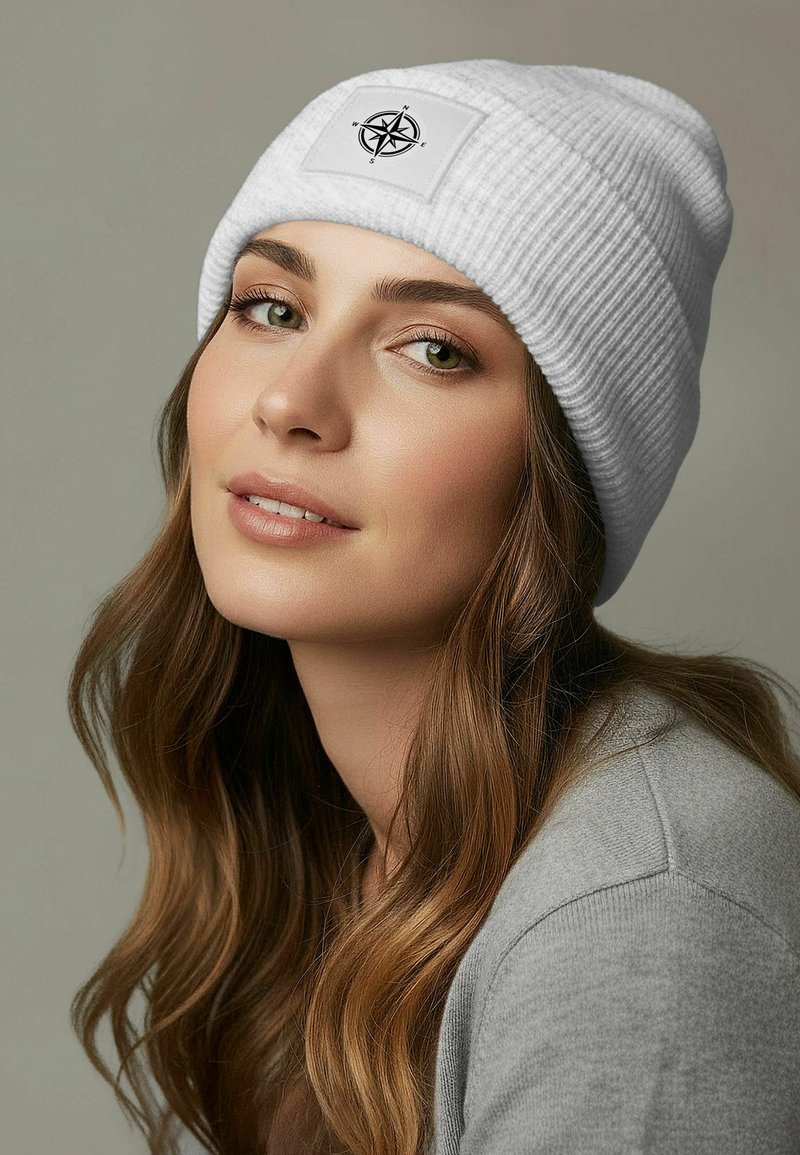 White ribbed beanie with a rectangular patch featuring a black compass design. Soft texture, fitted style, displayed on a neutral background.