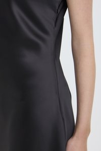 Fekete szatén ruha közeli képe, oldalról, a hónaljnál látható cipzárral, valamint egy meztelen kar világos háttér előtt.