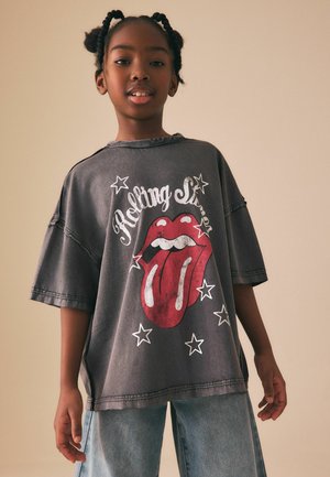 Młoda osoba z warkoczami nosi oversize szarą koszulkę Rolling Stones z czerwonym logo języka i jasnoniebieskie dżinsy na jednolitym tle.