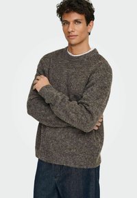 Pull brun texturé avec un col rond et des manches longues, associé à un jean en denim foncé. Le tissu semble doux et épais.