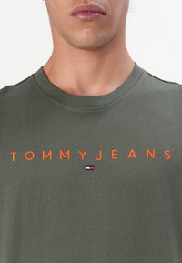 Groene katoenen t-shirt met oranje geborduurde "TOMMYJEANS" tekst en een klein vlaglogo op de borst. Ronde hals, korte mouwen.