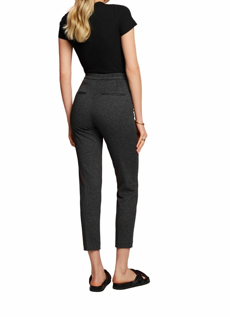 Žena s dlouhými blond vlasy, oblečená v černém tričku s krátkým rukávem, přiléhavých tmavě šedých zkrácených kalhotách, černých klouzavých sandálech a zlatých náramcích.