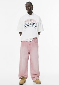 Przytulny, oversize'owy biały t-shirt z logo i grafiką, nałożony na koszulę w paski. Jasnoróżowe, szerokie spodnie dżinsowe i beżowe obuwie z sznurówkami.