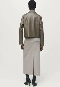 Une personne aux cheveux courts porte une veste en cuir olive raccourcie, une longue jupe beige avec une fente à l'arrière et des bottes à talons noires, se tenant de dos.