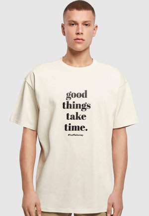 Beige bomull t-skjorte med et svart tekstgrafikk som skriver "gode ting tar tid." inkluderer hashtaggen "#TrustTheJourney."