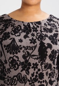 Motif floral noir sur un tissu gris avec un col froncé. Le design présente diverses fleurs et feuilles, créant un aspect texturé.