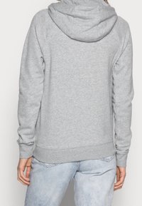 Personne portant un sweat à capuche gris clair et un jean bleu clair délavé, vu de dos contre un fond uni.