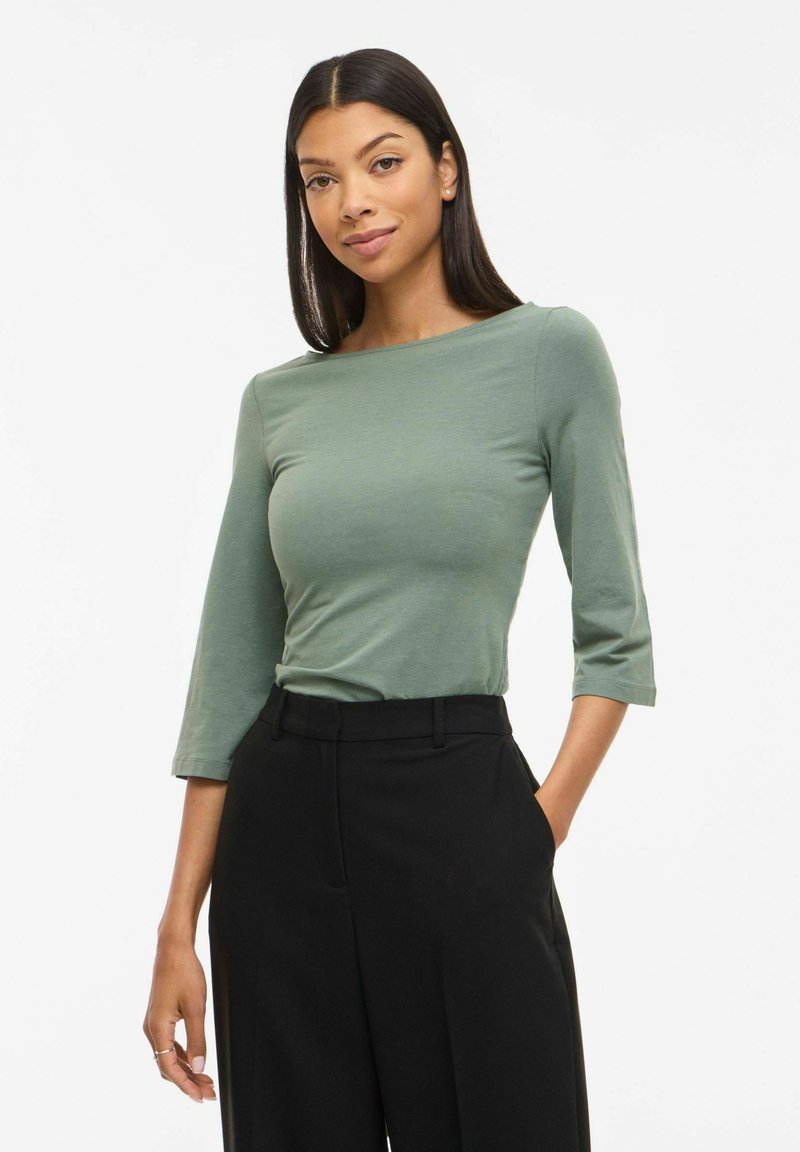 Junge Frau mit langen dunklen Haaren, die ein grünes Oberteil mit dreiviertellangen Ärmeln und eine schwarze hochtaillierte Hose trägt, stehend mit einer Hand in der Tasche.