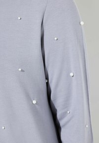 Top a maniche lunghe grigio chiaro con una texture morbida, caratterizzato da decorazioni in perle di dimensioni irregolari lungo la manica.