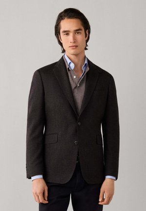 Jonge man draagt een donkerbruin blazer over een grijze trui en een blauw gestreept overhemd, staande tegen een effen lichtgrijze achtergrond.