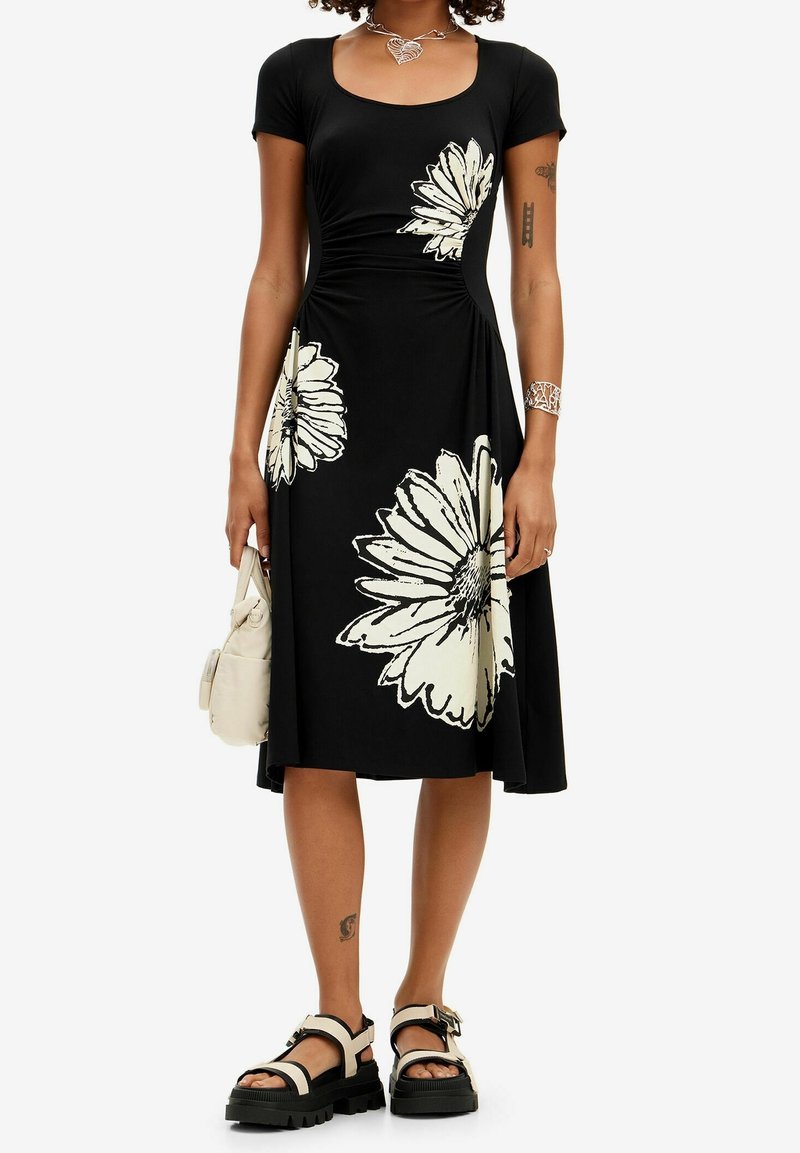 Femme portant une robe midi noire à grands imprimés floraux blancs, des sandales chunky noires et blanches, tenant un petit sac à main beige.