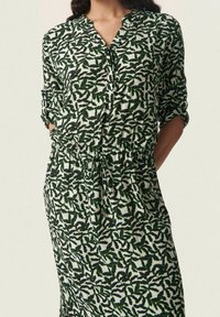 Vestido camisa de estampado de hojas en verde y negro, con mangas tres cuartos, escote en V y cintura fruncida. Tela ligera, corte relajado.