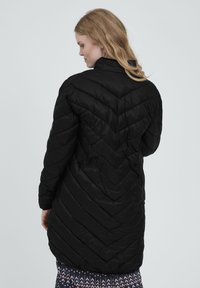 Fekete, steppelt hosszú kabát, chevron mintázattal, magas gallérral és oldalsó zsebekkel. Simma, fényes anyag, testhezálló siluettel.