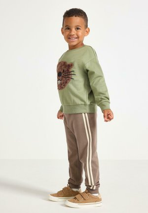 Zielony sweatshirt z brązową aplikacją bobra, w połączeniu z brązowymi spodniami dresowymi z białymi paskami oraz beżowymi trampkami na rzepy.