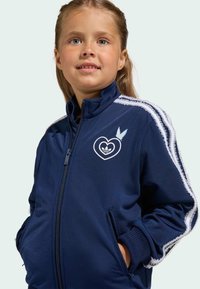 Námořnická modrá bunda na zip s bílými pruhy, logem srdce a detaily motýla. Obsahuje kapsy a žebrované manžety pro větší funkčnost.