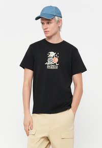 Black cotton t-shirt with a graphic print featuring a wave, globe, and accents in pastel colors. Model wears light denim cap and beige shorts.