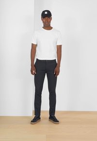 Camiseta blanca de manga corta, pantalones negros ajustados y zapatillas negras. Una gorra negra con un logo cuadrado completa el conjunto. El fondo es blanco liso.