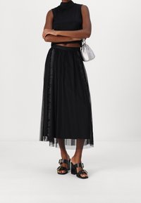Femme portant un top sans manches noir, une jupe midi noire transparente, des sandales à talons noires avec des boucles dorées, et tenant un petit sac à main en argent.