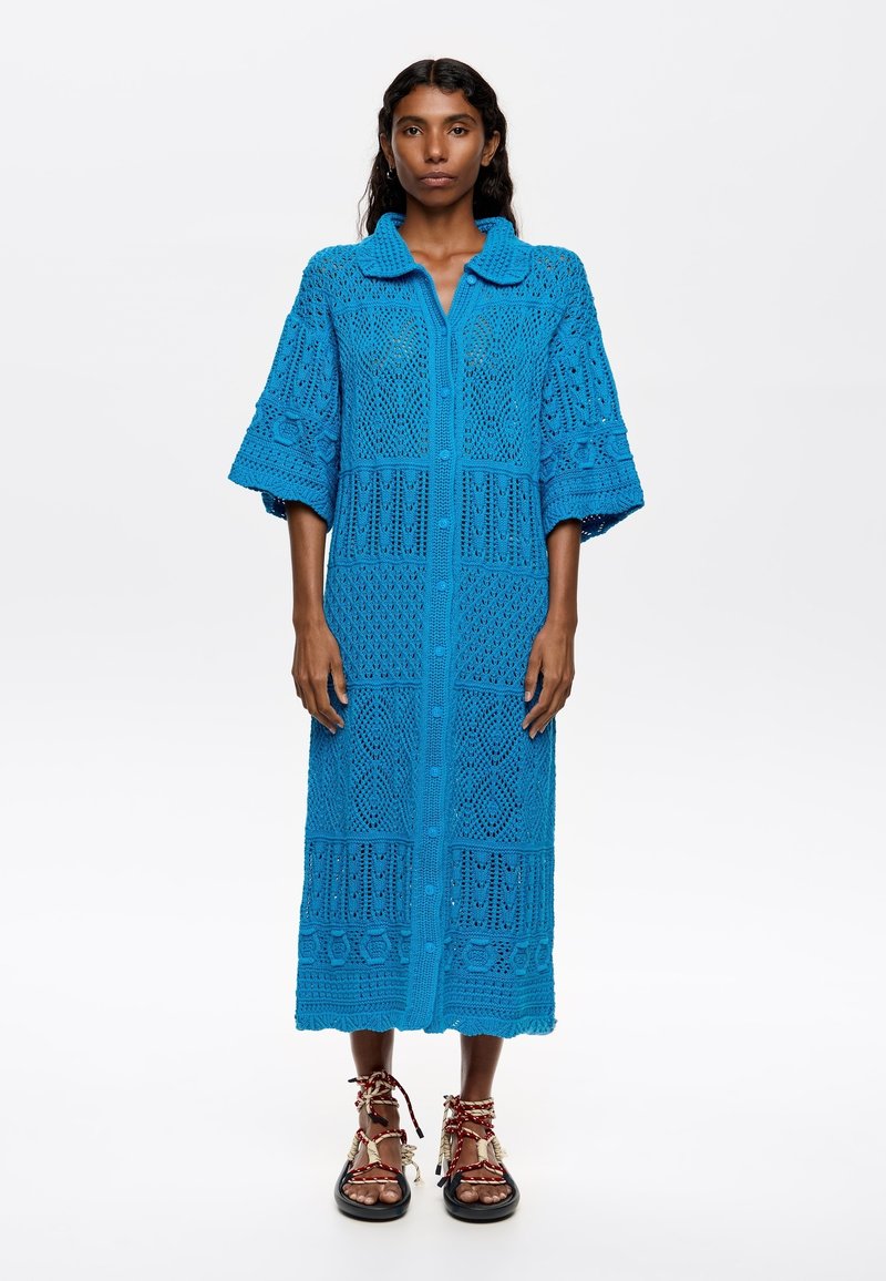 Abito blu lavorato a maglia con colletto, chiusura a bottoni, maniche ampie e intricati motivi in pizzo. Indossato con sandali dettagliati.