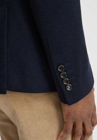 Zbliżenie na rękaw ciemnogranatowej marynarki z czterema czarnymi guzikami na tle jasnobrązowych spodni sztruksowych i ręki osoby.