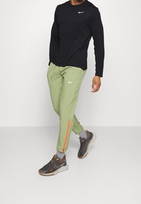 Schwarzes Langarm-Nike-Shirt, hellgrüne Hose mit orangefarbener Streifenverzierung, kombiniert mit dunklen Schuhen. Enthält Nike-Logos auf Shirt und Hose.