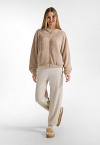 Chaqueta cremosa de felpa con cremallera y capucha, combinada con pantalones brillantes en un tono beige claro y zapatillas marrones. Textura suave en todo el conjunto.