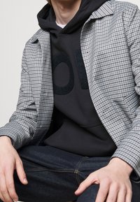 Chaqueta gris a cuadros con cuello solapa superpuesta a una sudadera con capucha gris oscuro que presenta un logo audaz y texturizado. Unos jeans oscuros completan el conjunto.
