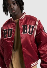 Röd satin varsity-jacka med svarta bokstäver "FU" och "BU", randiga ribbade muddar och broderad grafik på ärmen. Brun skjorta under.