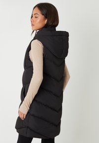 Frau trägt einen langen schwarzen, wattierten, ärmellosen Mantel mit Kapuze über einem beigen Langarmshirt, steht mit den Händen in den Taschen und blickt nach links.