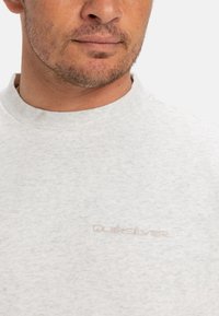 Světle šedé tričko s krátkým rukávem a kulatým výstřihem vyrobené z měkké tkaniny, s jemným texturovaným vzorem a malým vyšitým logem na hrudi.