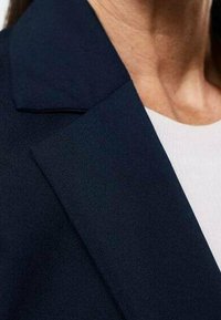 Blazer bleu marine avec un col structuré, présentant une texture lisse et une coupe ajustée, partiellement visible au-dessus d'un haut de couleur claire.