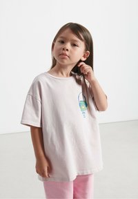 Šviesiai rožinė per didelė T-marškiniai su apvalia apykakle ir trumpomis rankovėmis. Priekyje yra mėlynos ir žalios spalvų grafika.